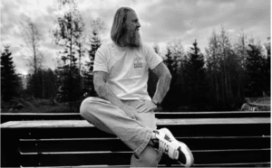 A bearded man with long hair sits on a wooden railing outdoors. He's wearing a printed t-shirt, jeans, and sneakers. The background shows trees and a cloudy sky. The image is in black and white.